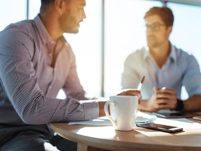 Closeup of coffee cup with blurred image of two businessmen sitting on table.