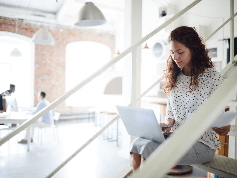 Young businesswoman sitting on steps of staircase with laptop on her knees and searching for information in the net.