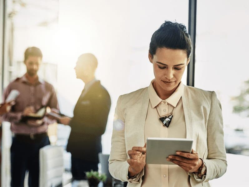 Businesswoman using her tablet while some colleagues talk in the background.