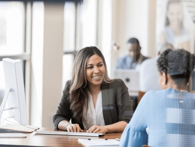 Happy female bank employee uses a desktop computer while assisting a female customer with a new account.