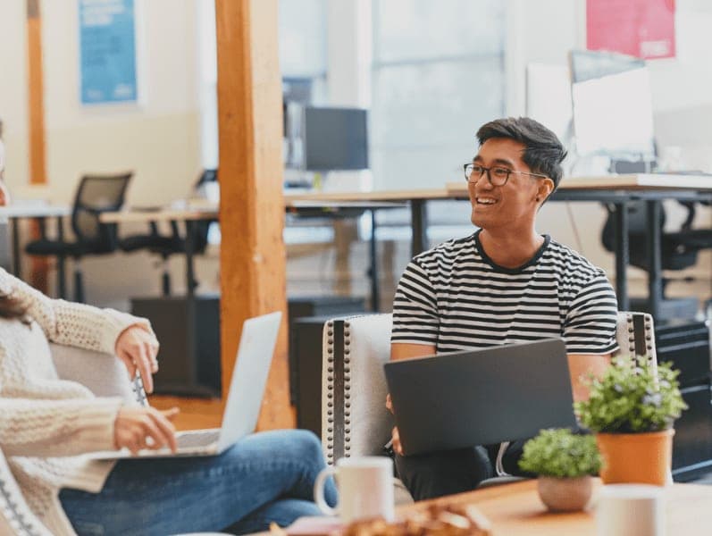 Smiling male employee sitting, holding laptop talking with coworker in the office.