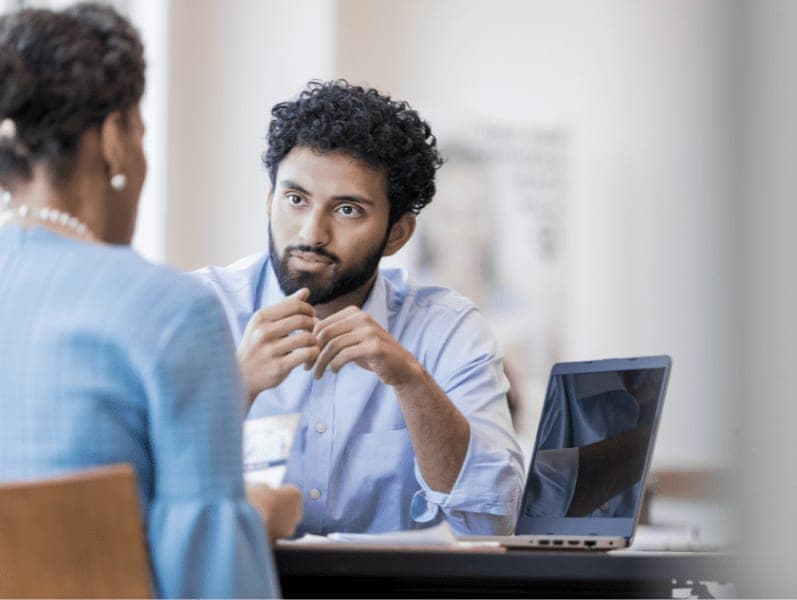 A young male accountant sits and makes eye contact with his senior female client as he listens to her raise concerns about her firm.