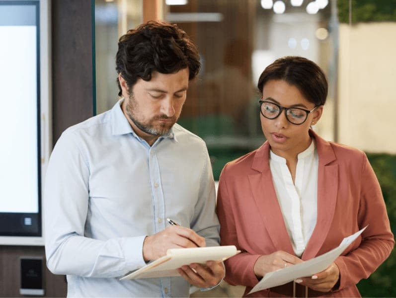 Two business colleagues checking their notes and comparing the business plan while standing at office.