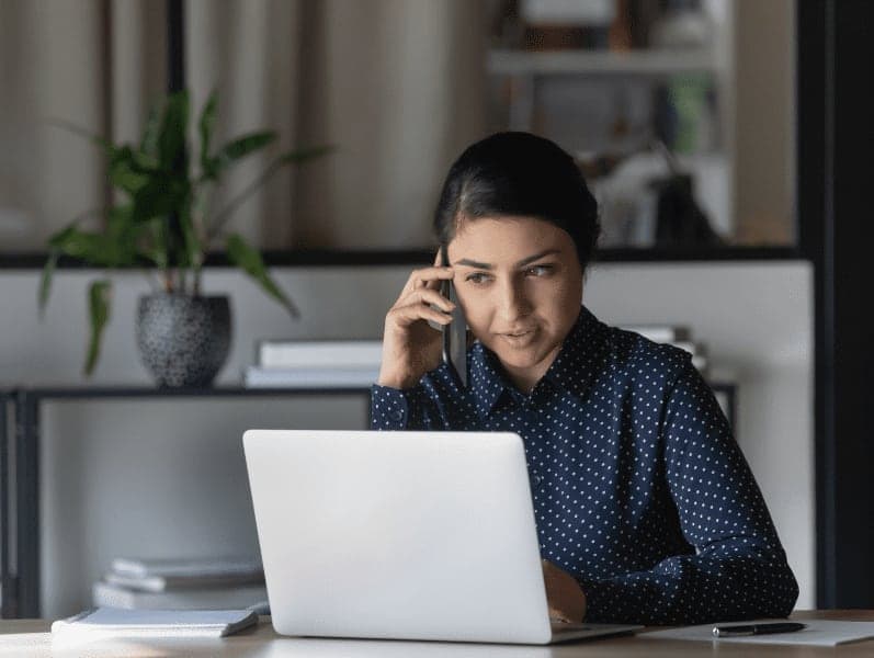 Businesswoman sitting at table with laptop, having a phone call with client in office.