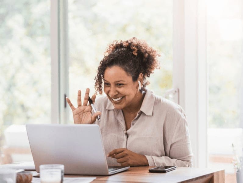 Businesswoman waving hello to coworkers on laptop during virtual meeting.