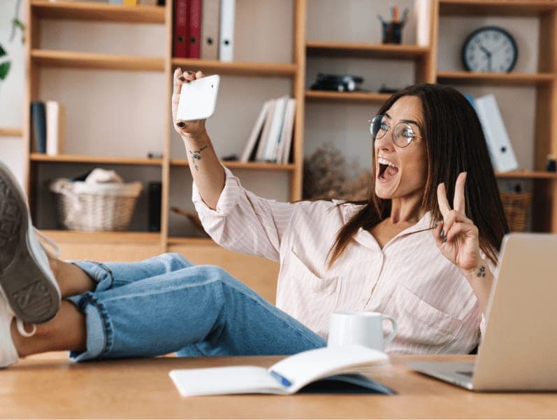 Positive happy young business woman in office taking a selfie by mobile phone and using laptop computer