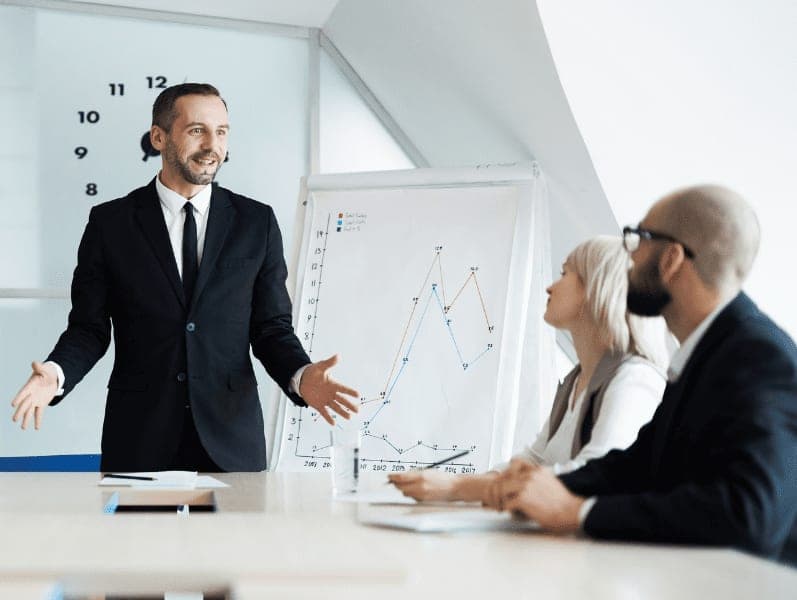 Confident business coach standing by table in front of employees and explaining his principles of successful work organization
