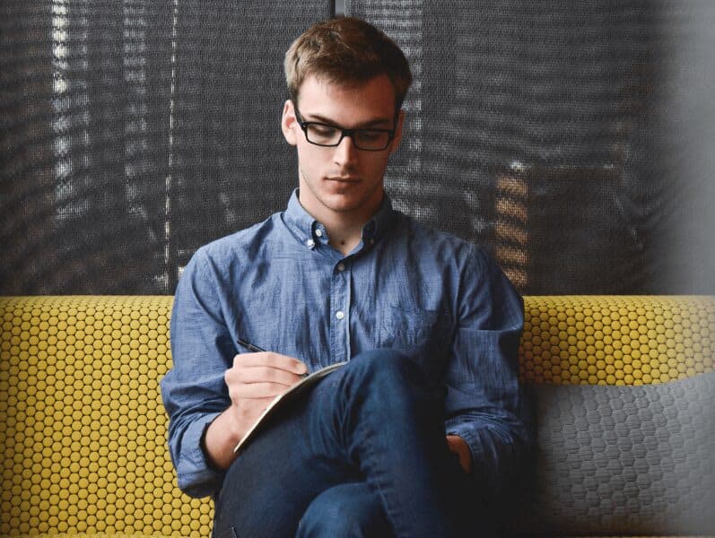 Younger male sitting on yellow couch with glasses with legs crossed writing on a book
