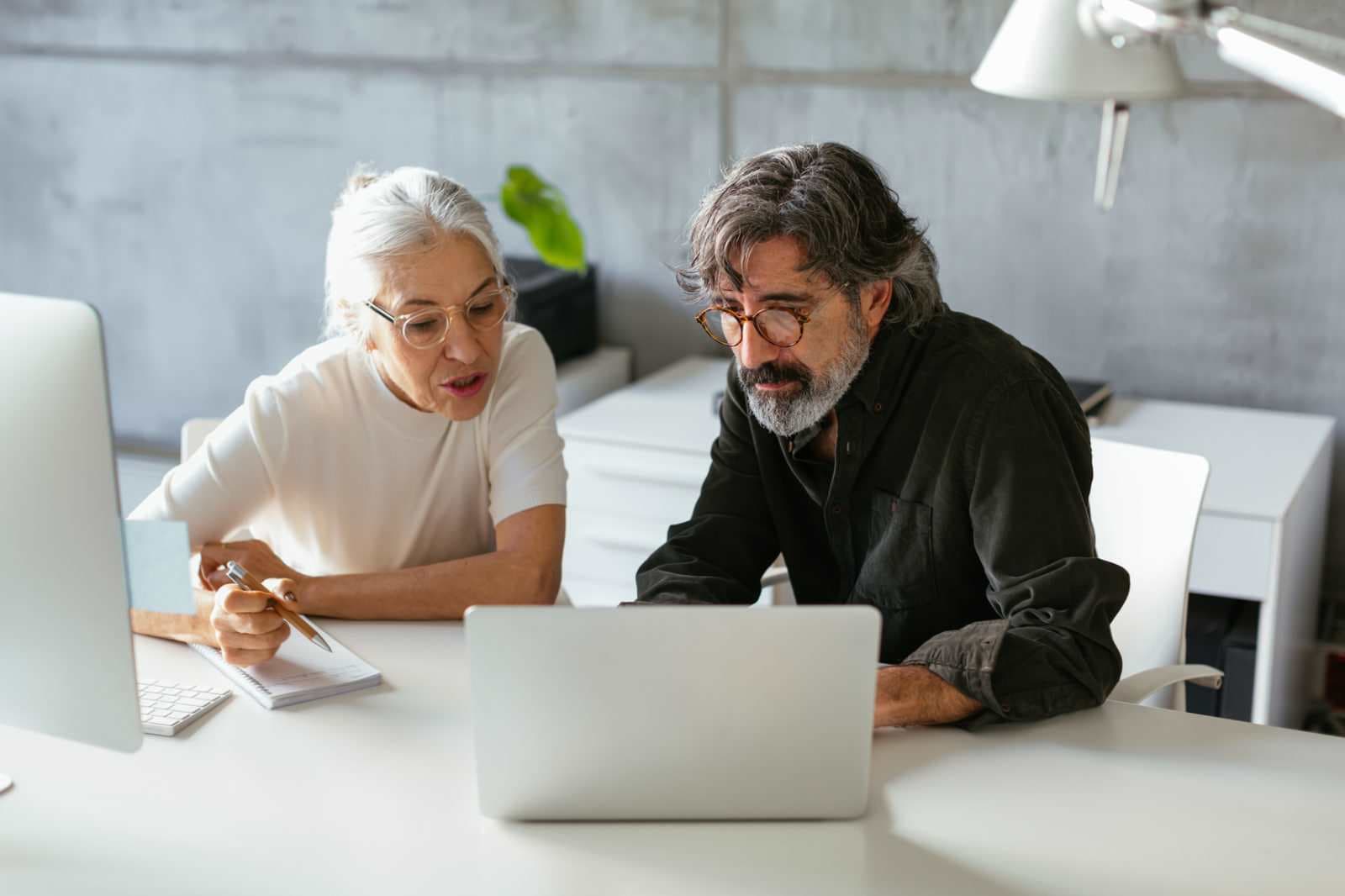 Serious mature woman making notes and explaining data to bearded male coworker while using laptop in modern office together