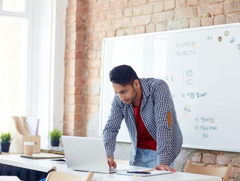 Young male business owner standing over desk and lap top review items on the screen with a white board behind him