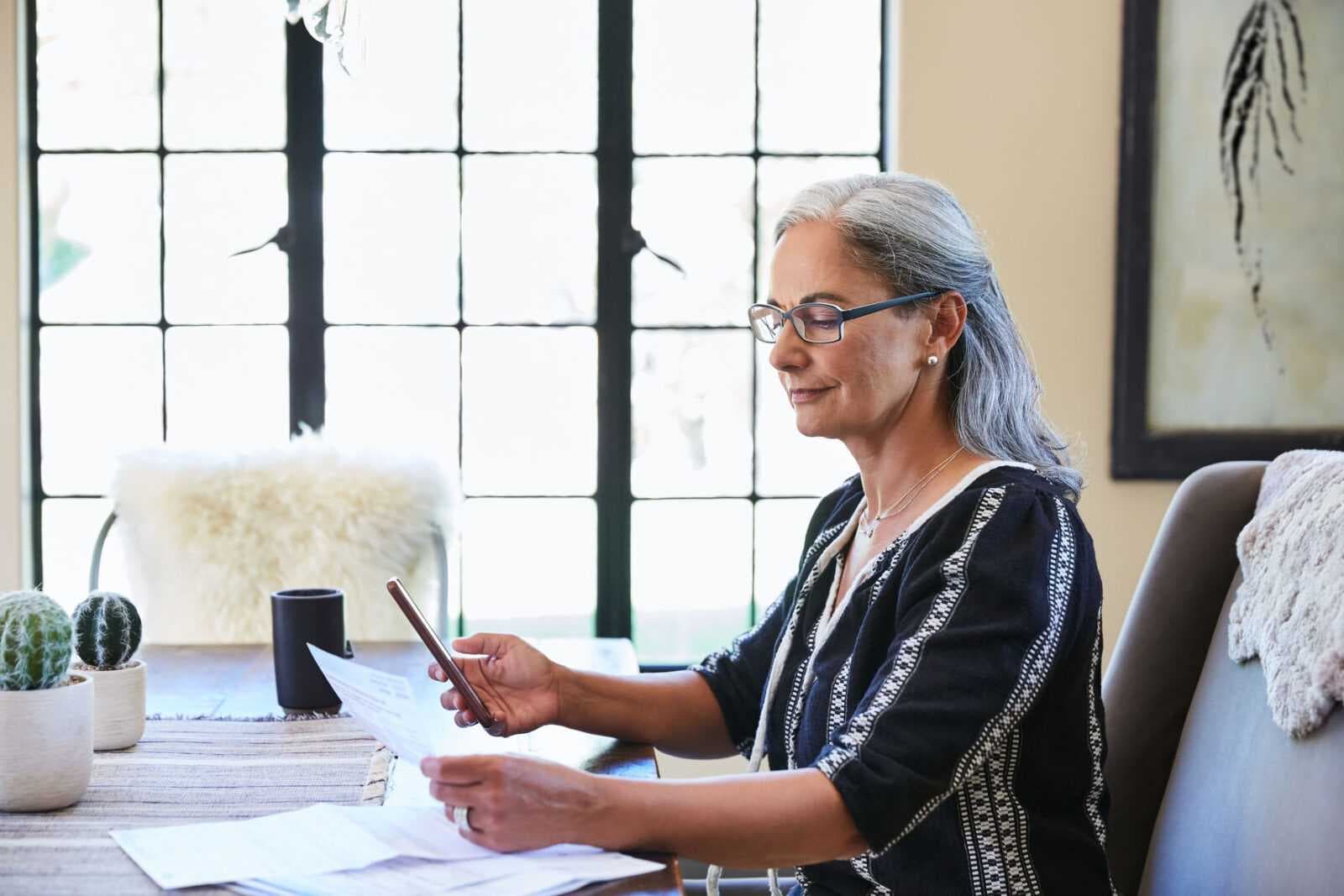 Mature woman with grey hair at home paying bills on her mobile phone at dining room table