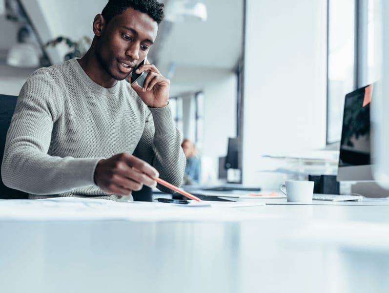 Man sitting at his desk on his cell phone referencing a document in front of him