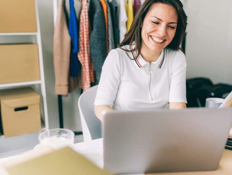 Woman siting at desk with clothing behind smiling while working on her computer