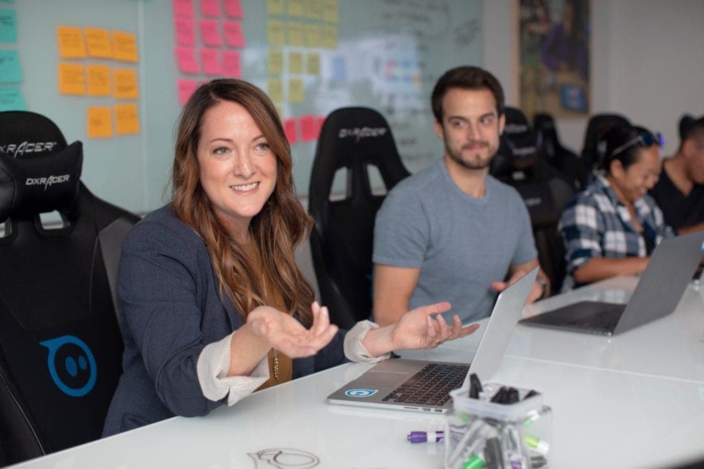Woman extending her hand as is speaking while sitting at a desk beside colleagues
