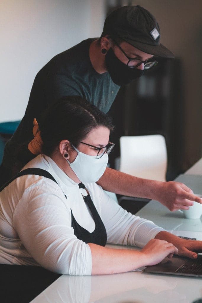 A woman siting and working at a table with a standing beside her supporting her while both wearing masks