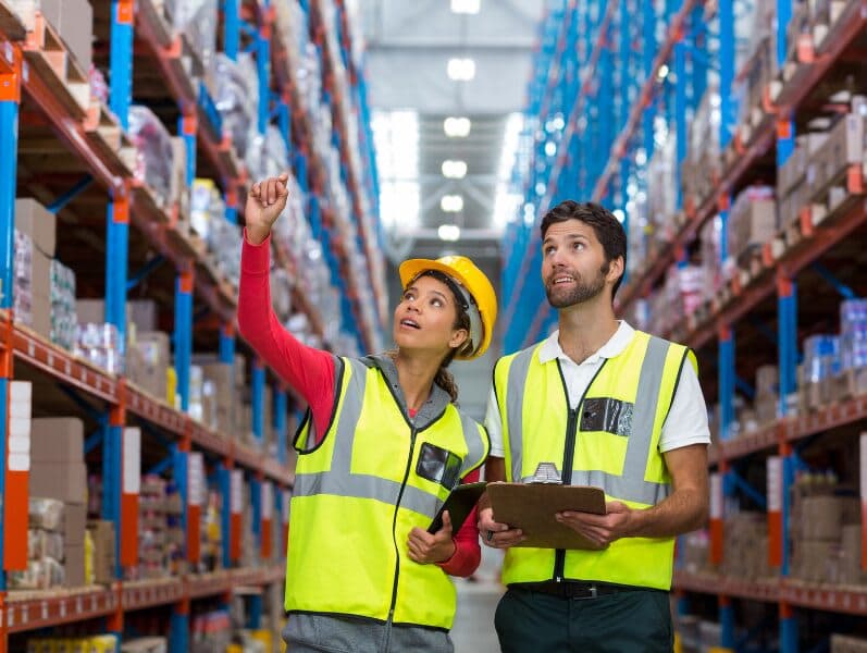 Male and female employee in warehouse looking up wearing safety gear