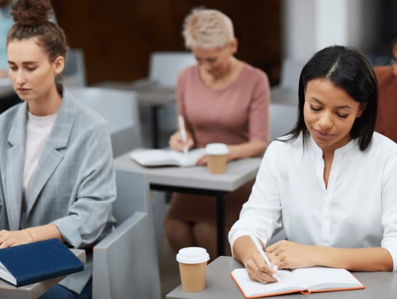 Businesswoman writing notes while sitting at table in class with group of students.