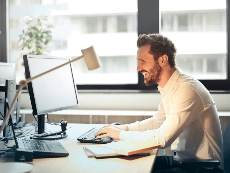 Man in white shirt sitting on chair while facing black computer set and smiling.