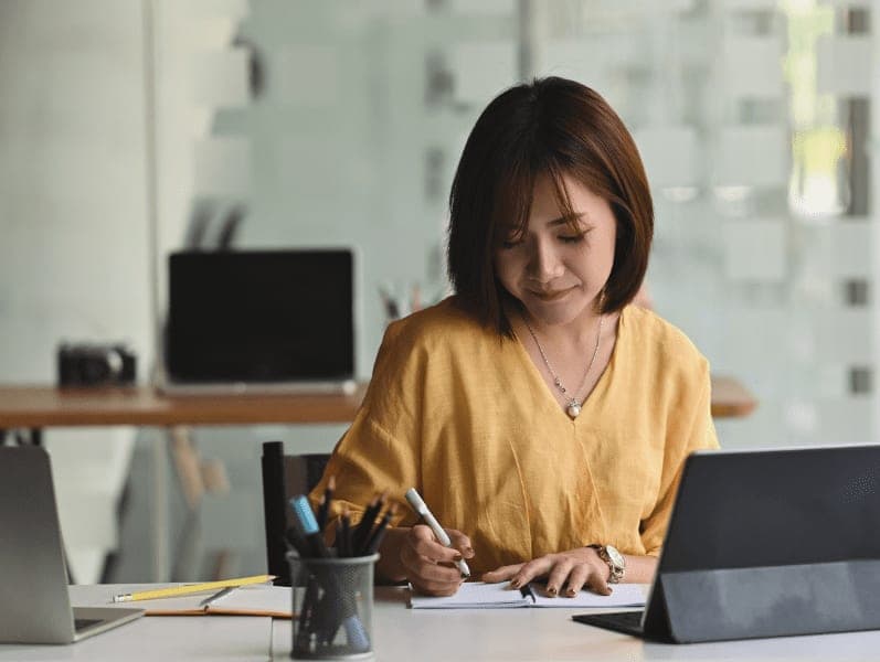Young woman writing on paper while sitting in front her computer tablet in a modern office