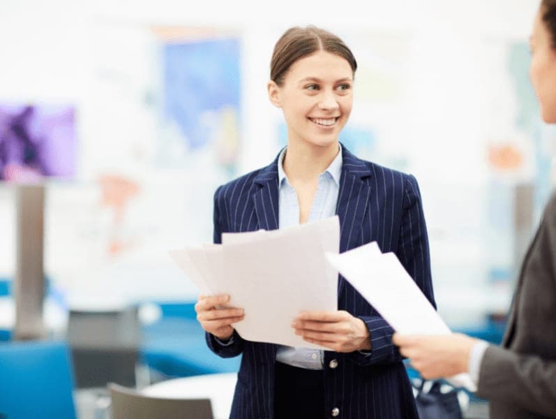 Smiling young businesswoman talking to colleague while standing in office at work