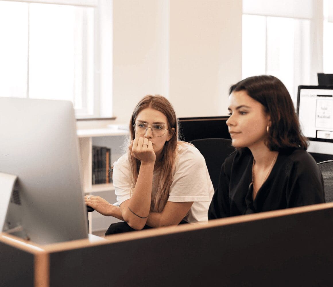 Two women looking at a desktop monitor together at a shared workstation in a bright office