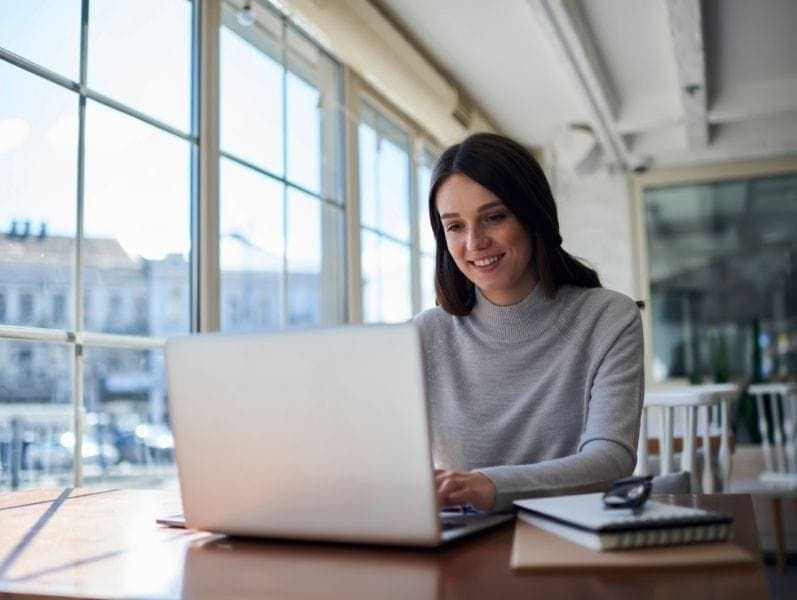 Female freelancer sitting in front of desk with not pad and lap top by windows in an open space area