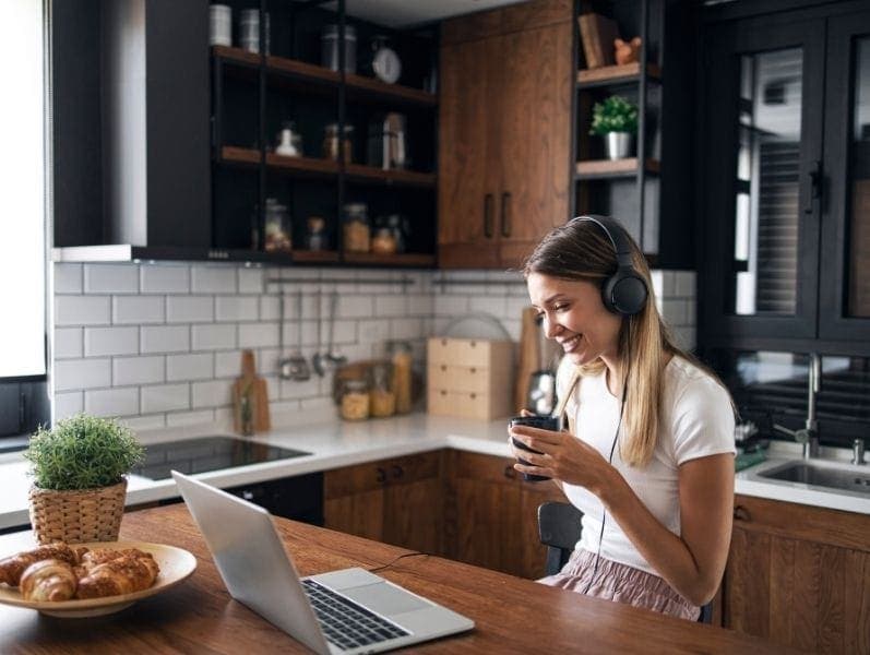 Young female freelancer sitting in her kitchen in front her laptop with head phones on holding a drink