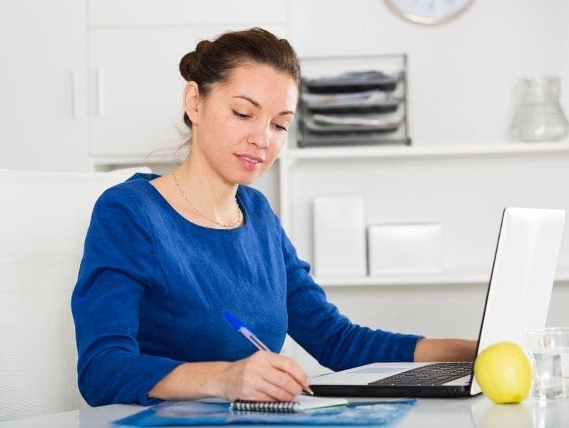 Woman sitting in front of desktop computer writing notes on a note page to the side of her