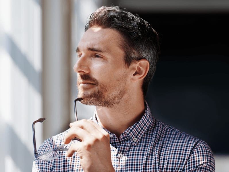 Man looking out the window holding his glasses thinking about self-awareness