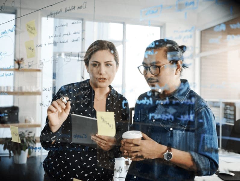 Male and female employee standing in front of glass white board reviewing items