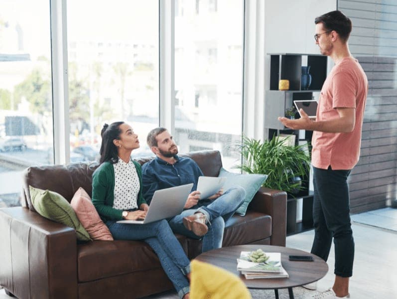 Two colleagues sitting on a couch facing a male colleague standing and speaking