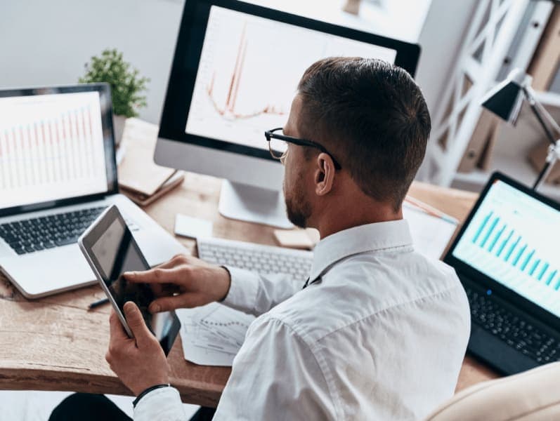 Rear view of man sitting in front of desk with 3 screens and a table in front of him