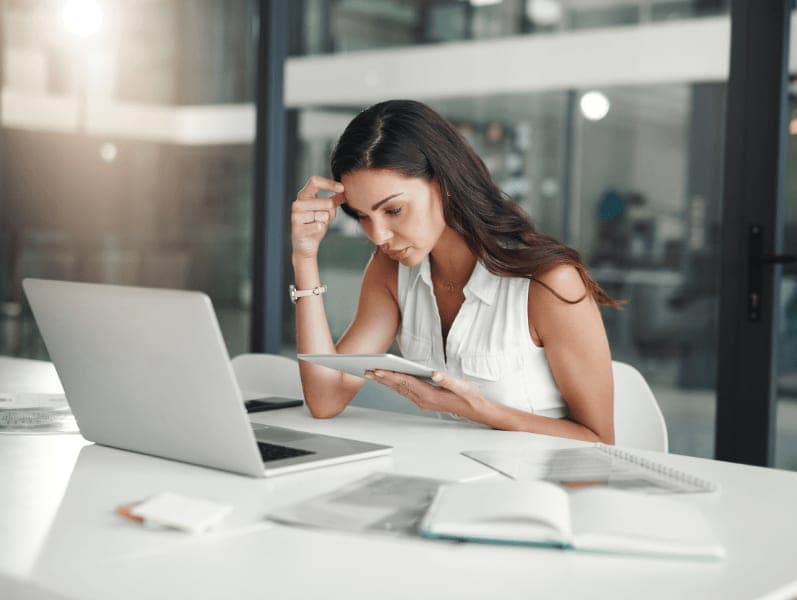 Woman sitting in glass office area referencing tablet and laptop