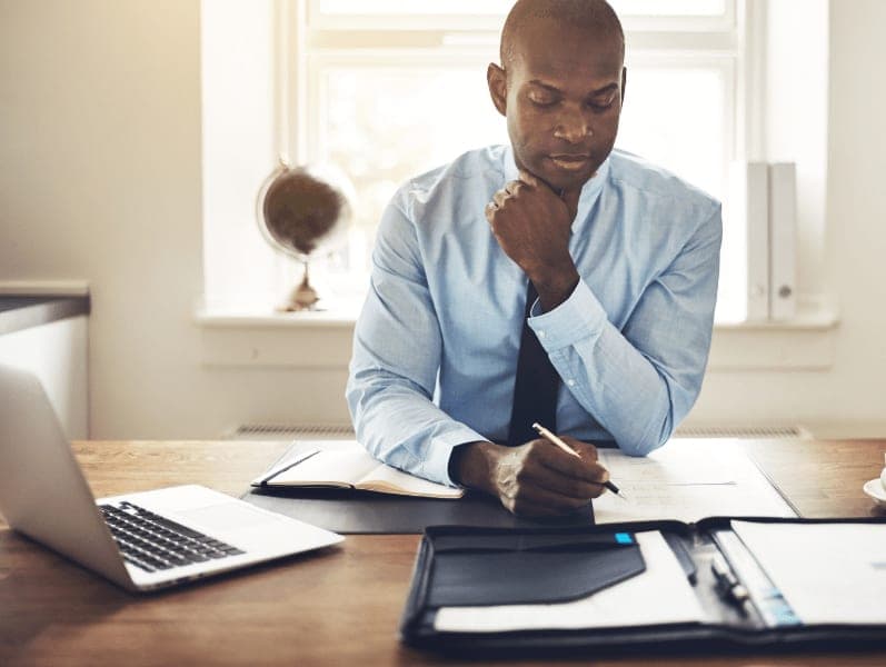 Man sitting front view at desk with binder open and laptop to the side of him writing on a document