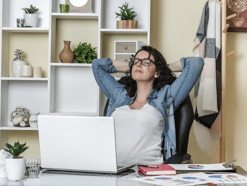 Female sitting at desk with laptop in front of her with hands resting behind head with eyes closed