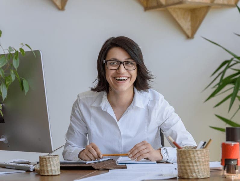 Woman sitting at desk with paper work in front holding a pen smiling