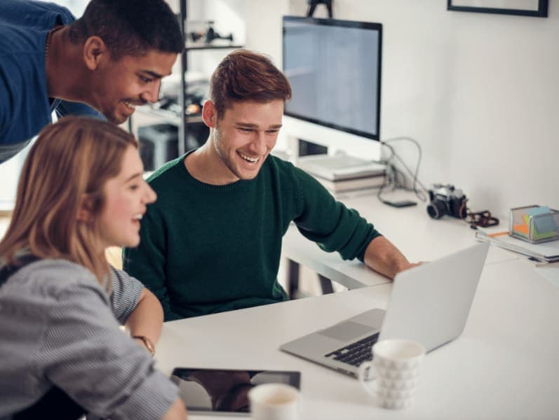 two young employees sitting at a desk with a one standing looking over to view what is on the lap top smiling