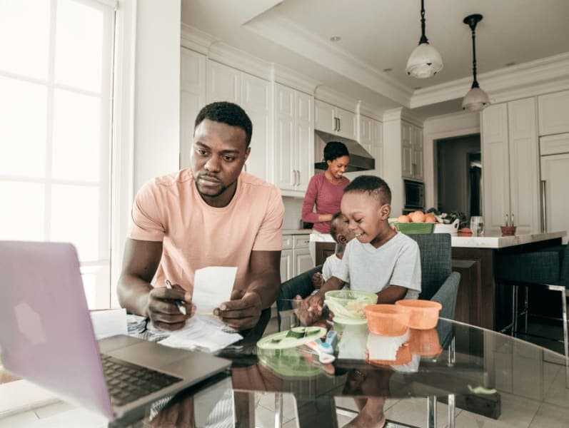 Man sitting at kitchen table with young kid next to him referencing his laptop