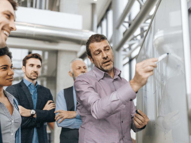 Senior male colleague writing on white board surround bay four employees