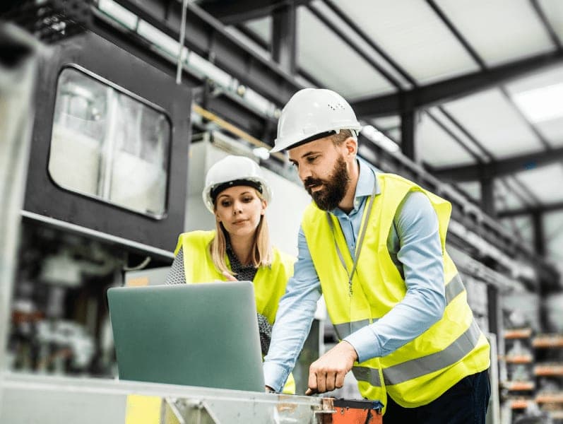 Male and female wearing safety vest and hard hats disusing something over a laptop while in manufacturing facility