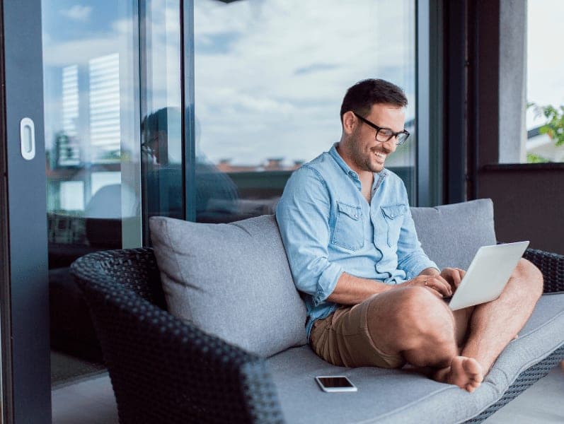 Man sitting outside on patio furniture with legs crossed with laptop on his lap