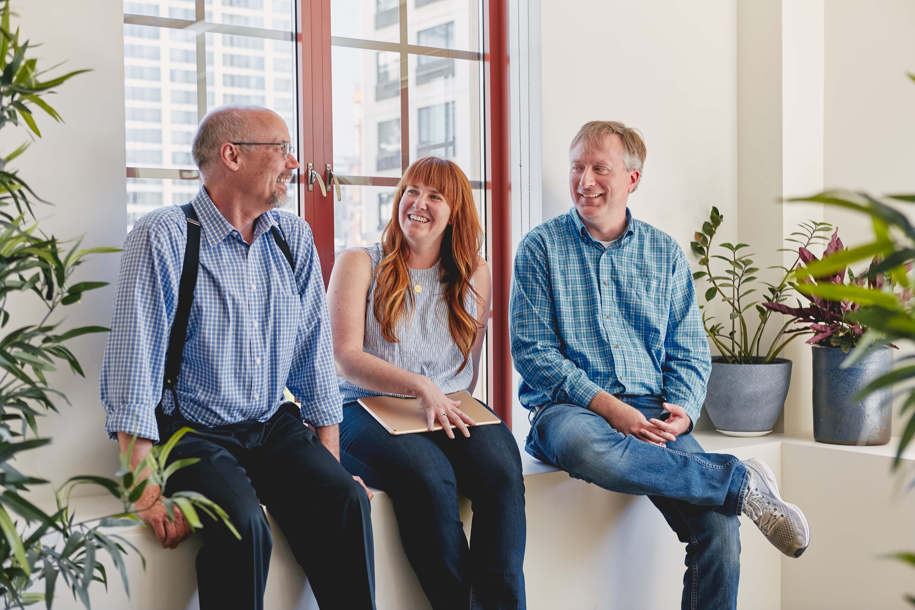 Three colleagues laughing and talking together while seated on a window ledge in a bright, plant-filled office with a city skyline in the background