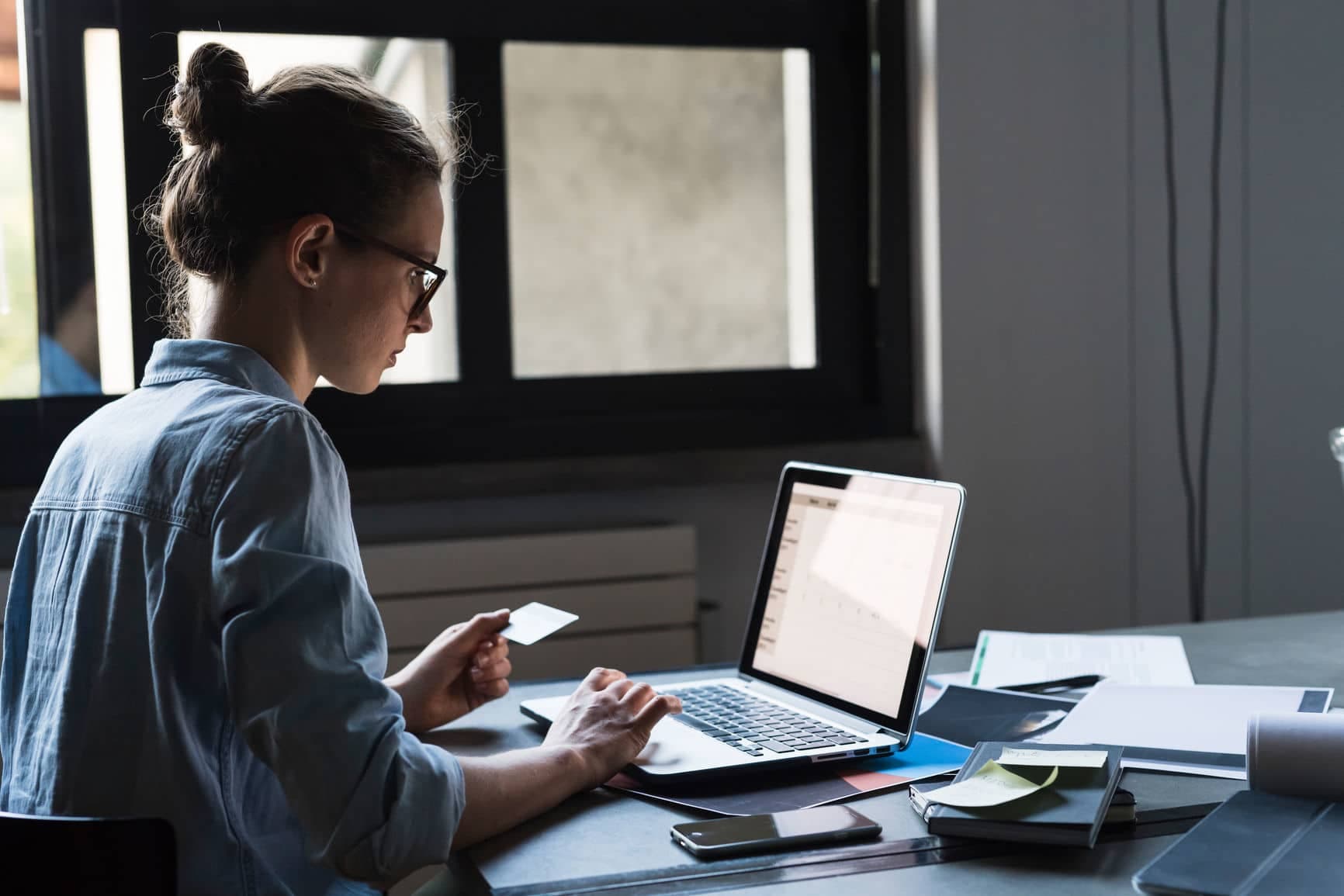 Woman in glasses holding a card while typing on a laptop showing a form or spreadsheet at her desk