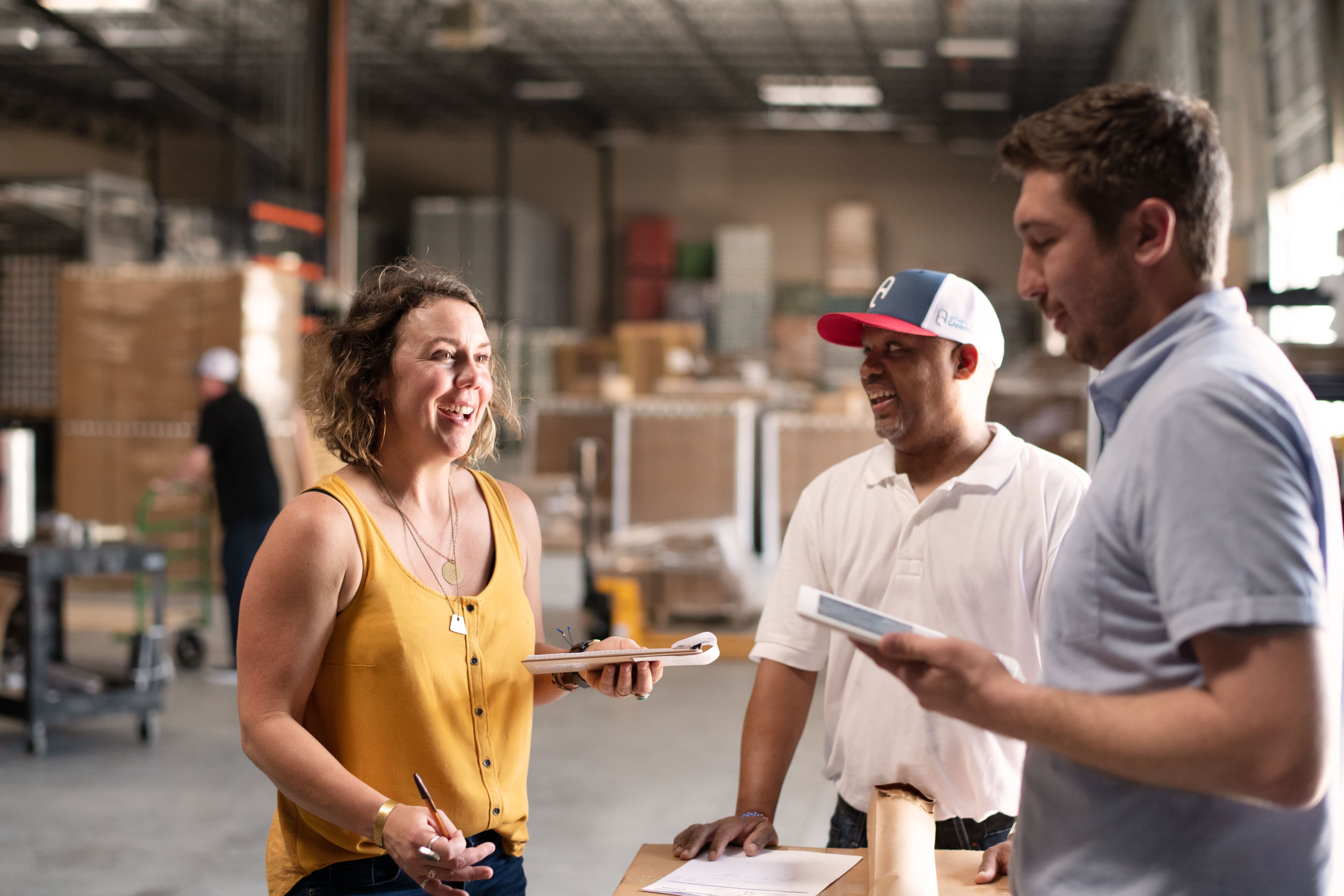Three coworkers talking and smiling on a warehouse floor, holding clipboards and paperwork, with stocked shelves behind them
